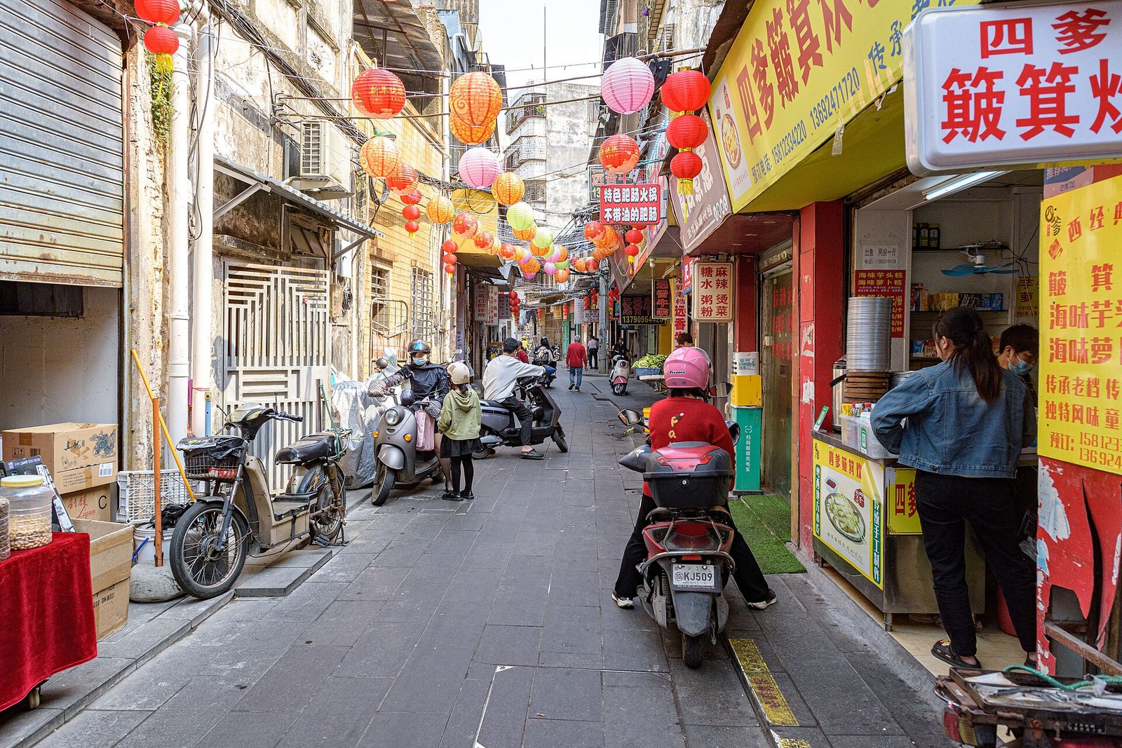 A narrow alley strung with paper lanterns, lined with food stalls and parked electric scooters, with residents walking and a woman working at a steamer counter