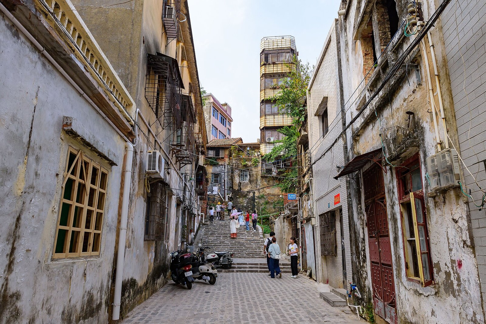 A stone-paved lane climbing a flight of steps between weathered two-storey buildings, with residents walking and scooters parked at the bottom
