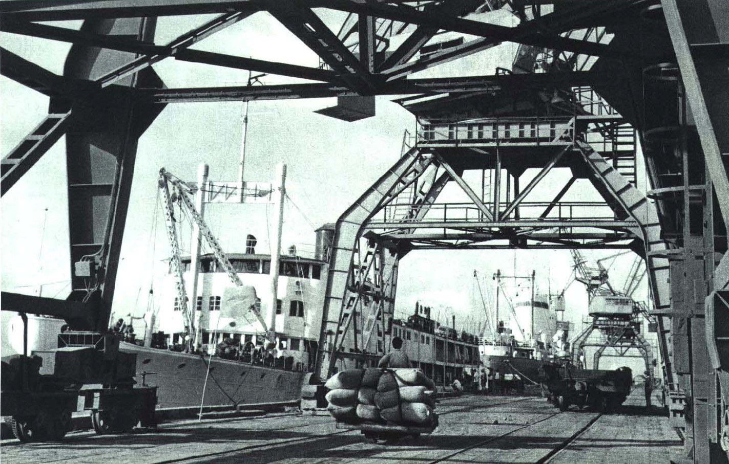 A black and white photograph of a busy port with cargo ships, cranes and warehouses, dated 1965