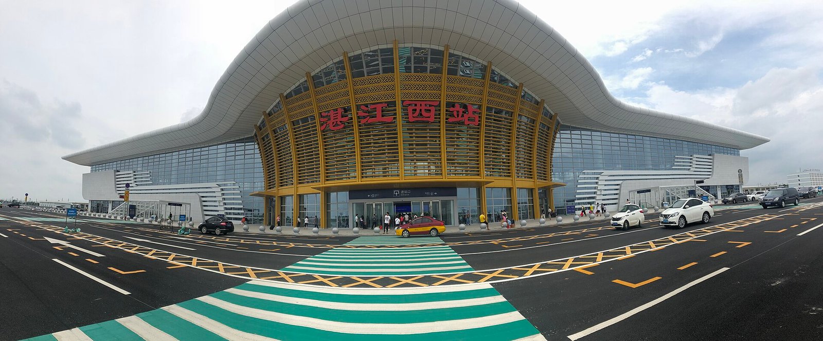 A wide panoramic view of a modern railway station with a sweeping curved roof and a golden lattice facade, with a striped pedestrian crossing in the foreground
