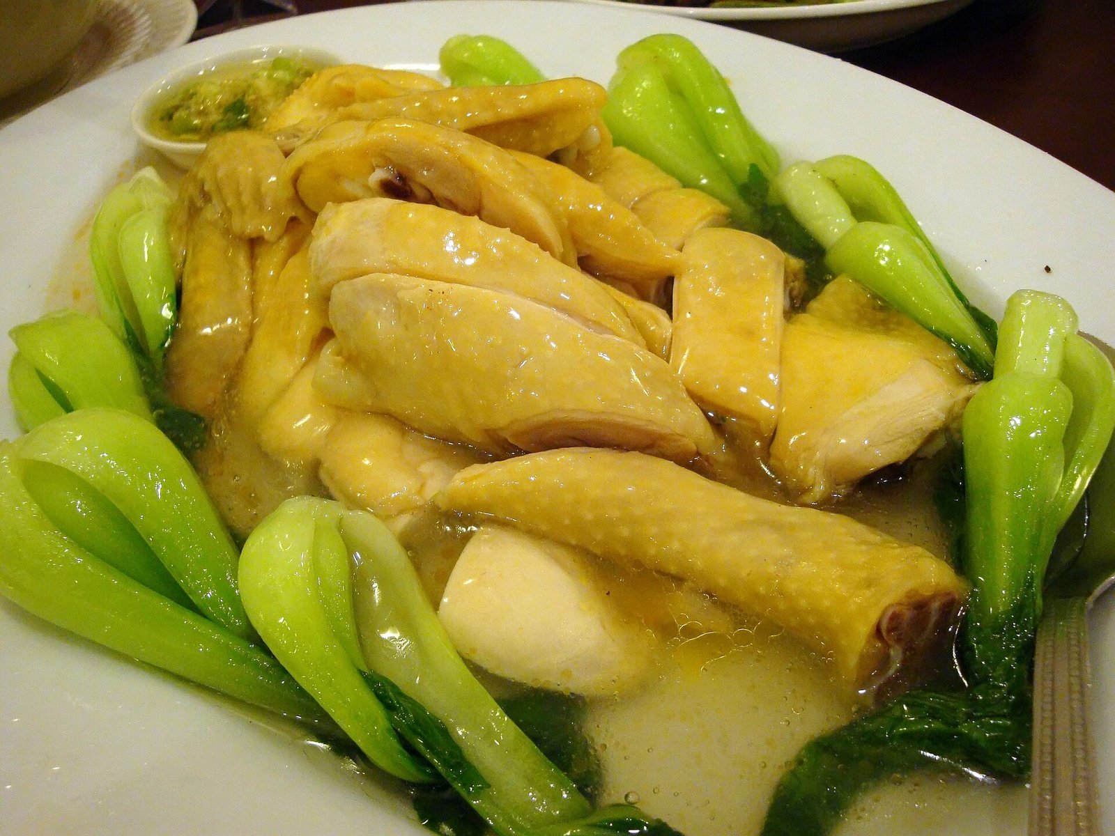 A plate of pale poached chicken cut into pieces, skin intact and glistening, garnished with green leaves