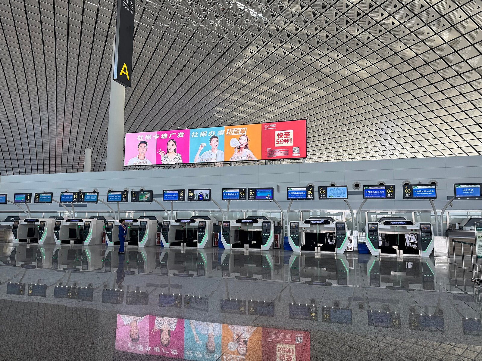 A wide, almost empty airport check-in hall with a row of self-service kiosks, a large LED billboard and a highly reflective floor