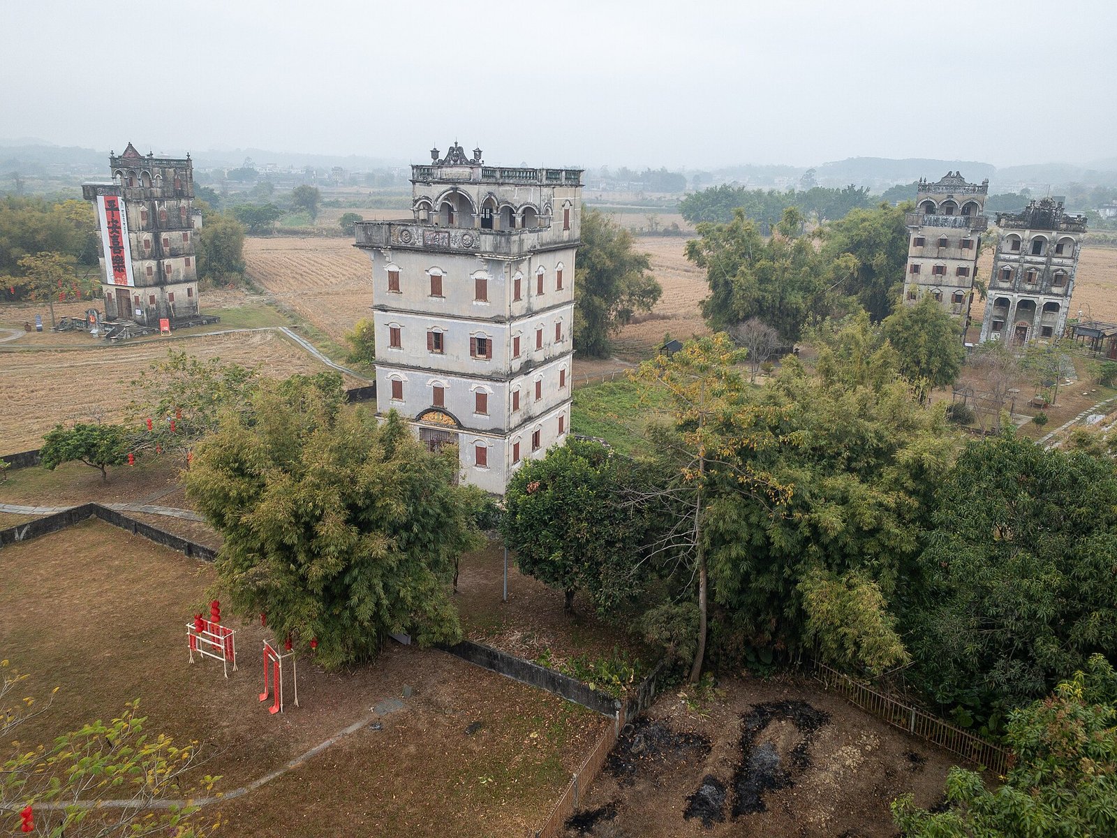 A village of grey tiled roofs and fortified towers seen from the rooftop of a taller tower, with fields beyond